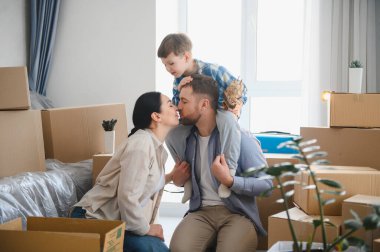 Genç bir aile yeni evlerine taşınıyor, ebeveynler babalarının omuzlarında çocukla öpüşüyor, etrafı karton kutularla çevrili.