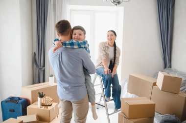 Çocukları kucaklayan, gülümseyen ve mukavva kutularla çevrili aileler mutlu mesut yeni evlerine taşınıyorlar.