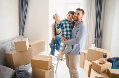 Üç kişilik mutlu bir aile taşındıktan sonra karton kutularla çevrili yeni bir evde kucaklaşıyor, gülümsüyor ve başparmak kaldırıyor.