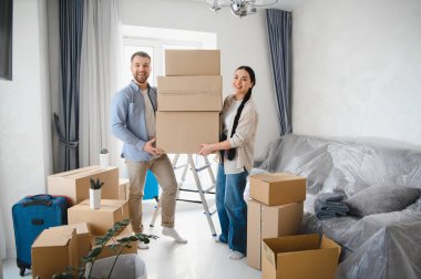 Genç çift gülümsüyor, birlikte karton kutular taşıyorlar, yeni bir eve taşınıyorlar. Onları çevreleyen oda nakliye malzemeleri ile dolu.