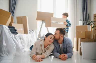 Mutlu bir aile yeni bir eve taşınıyor. Ebeveynler yerde dinleniyor, baba anneyi alnından öpüyor, çocuk karton kutu taşıyor.