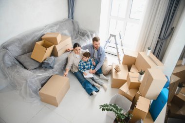Aile ve çocuk, karton kutuların arasında oturmuş taşınma günü için heyecanlı bir şekilde yeni bir apartman dairesinde geleceklerini planlıyorlar.