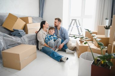 Genç bir aile mutlu ve yeni dairede karton kutuların arasında oturuyor, taşınıyor ve ev hayallerini gerçekleştiriyor.