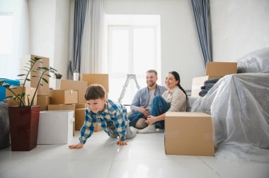 Neşeli ebeveynler ve oğulları karton kutularla dolu bir salonda yerde oynuyorlar ve yeni evlerine taşınıyorlar.