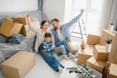 Mutlu ebeveyn ve çocuk ailesi, karton kutularla çevrili yerde oturuyor ve başarılı ev taşınmalarını kutluyorlar.