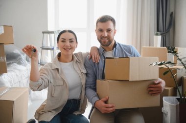 Heyecanlı çift apartmana taşınmayı kutluyor, kadın evin anahtarlarını gösteriyor, adam karton kutuları taşıyor.