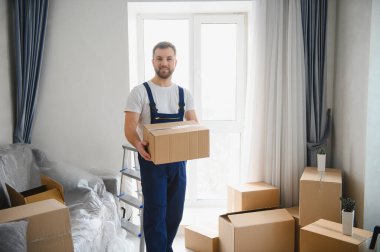 Üniformalı bir adam bir kutu taşıyor, yeni bir eve taşınırken mutlu hissediyor, etrafı eşyalarla çevrili.