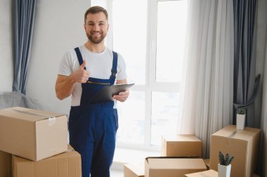 Üniformalı, gülümseyen, baş parmağını kaldıran, elinde bir pano tutan, karton kutularla çevrili yeni bir evde duran.