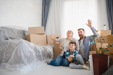 Genç bir aile, anne-baba ve oğul, yeni evlerini mutlu bir şekilde kutluyorlar, taşınan kutular ve mobilyalarla çevrili olarak yerde oturuyorlar.