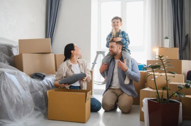 Aile, yeni dairelerinde eşyalarını boşaltıyor. Baba oğlunu sırtında taşırken anne kutuları düzenliyor.
