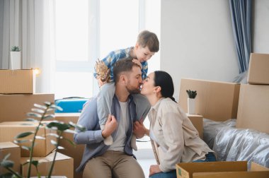 Mutlu aile yeni bir dairede karton kutuları açıyor. Ebeveynler babalarının omuzlarında çocuklarıyla öpüşürler.