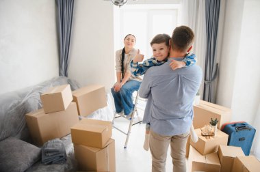 Mutlu bir aile yeni bir eve taşınıyor, çocuk onu kucağına alırken, anne yanında karton kutularla oturuyor.