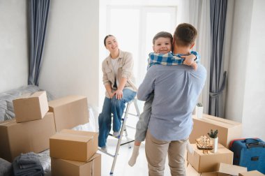 Genç bir aile taşınmayı ve yeni bir başlangıcı kutluyor. Karton kutuları açıyor, baba oğlunu kucağına alıyor ve anne mutlu bir şekilde izliyor.