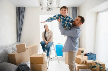 Üç kişilik gülümseyen aile, baba oğlunu tutarken anne izliyor, yeni dairede kutular taşınıyor.