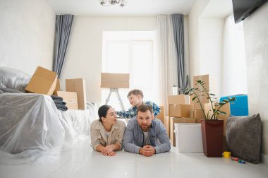 Mutlu ebeveynler ve çocuklar yeni evlerine taşındıktan sonra karton kutularla çevrili olarak yerde dinleniyorlar.