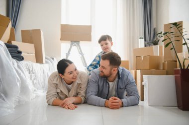 Ebeveynler ve oğulları karton kutularla çevrili bir şekilde yerde yatıyorlar ve yeni evlerine taşınmanın keyfini çıkarıyorlar.