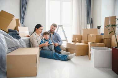 Karton kutuların arasında yerde oturan genç bir aile yeni bir dairede yerleşmeye ve yerleşmeye başlıyor.