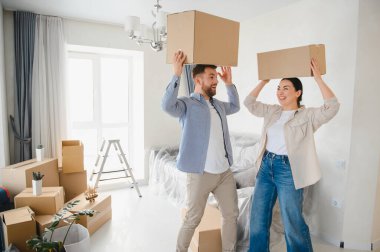 Genç adam ve kadın sevinçle taşınmayı kutluyorlar, kafalarında karton kutular taşırken yeni evlerini döşüyorlar.