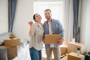 Genç çift taşınma gününü kutluyorlar, ellerinde anahtarlar ve karton kutular ile yeni evlerinde paketlenmemiş kutularla.