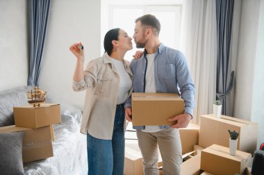 Mutlu çift yeni ev sahipliğini kutluyor, ellerinde dairenin anahtarları ve taşınma kutusu var, karton kutuların arasında duruyorlar.