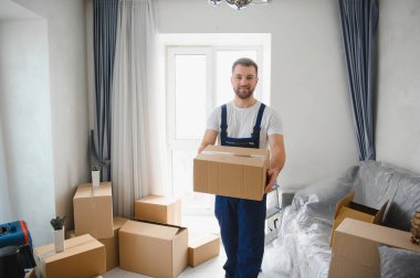 Üniformalı, elinde karton bir kutu, taşınma sırasında paketlenmiş kutularla dolu bir dairede çalışıyor.