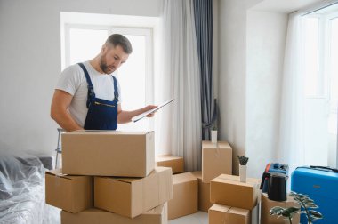 Üniformalı bir erkek taşıyıcı, taşınma gününde parlak bir dairede karton kutular ile birlikte bir pano üzerinde nesneleri kontrol ediyor.