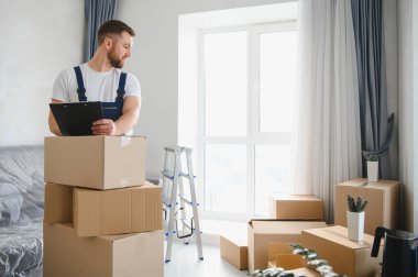 Profesyonel nakliyeci boş bir odada duruyor, karton kutularla çevrili bir panoda envanter listesini kontrol ediyor.