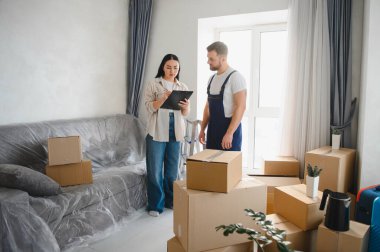 Genç bir kadın, profesyonel bir nakliyeci, kutular ve yeni bir dairede etraflarını saran mobilyalarla kontrol listesi imzalıyor.