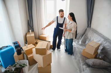 Profesyonel nakliyeci ve müvekkil karton kutular, bavullar ve örtülü bir kanepe ile dolu bir oturma odasında detayları tartışıyorlar.