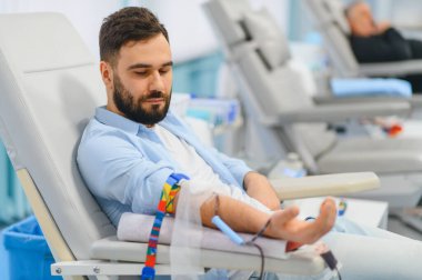 Genç bir adam kan bağışı yapıyor, tıbbi bir sandalyede oturuyor, sağlık hizmetlerine ve hayat kurtarma çabalarına katkıda bulunuyor.