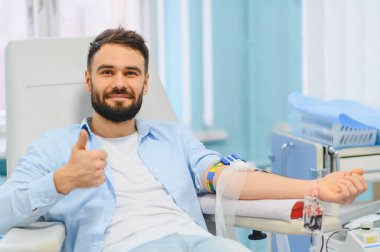 Genç adam gülümseyip başparmağını kaldırırken klinikte kan bağışlıyor, sağlık ve fedakarlığı destekliyor.