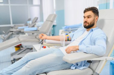 Klinik koltuğunda oturan genç adam kan bağışlıyor, tıbbi nakil sırasında hayat kurtarmaya odaklanmış.
