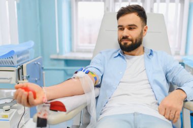 Mavi gömlekli ve beyaz tişörtlü genç bir adam bir sandalyede oturuyor, kan bağışlıyor, elinde kırmızı bir top tutuyor.