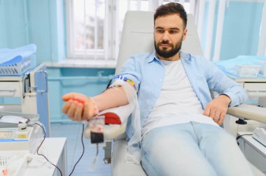 Adam klinik koltuğunda oturuyor, kan bağışlıyor, stres topu tutuyor, sağlık ve hayır işlerine katkıda bulunuyor.