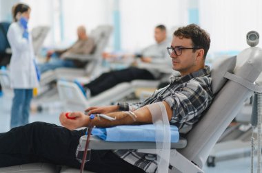 Kan bağışı yapan gözlüklü bir adam, arka planda bir hemşire ve diğer bağışçılarla birlikte yaslanmış bir sağlık koltuğunda oturuyor.