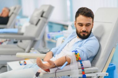 Genç adam klinik koltuğunda oturuyor, özverili bir kan bağışı yapıyor, sağlık hizmetlerini destekliyor ve ihtiyacı olanlara yardım ediyor.