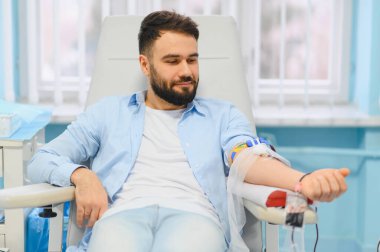 Sakallı ve mavi gömlekli bir adam klinikte kan bağışı yapıyor, insanlara yardım ediyor ve cömertçe hayat kurtarıyor.