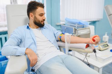 Sakallı bir adam kan bağışlıyor, kırmızı bir top sıkıyor, kan nakli için ekipmana bağlı olduğunu gösteriyor.