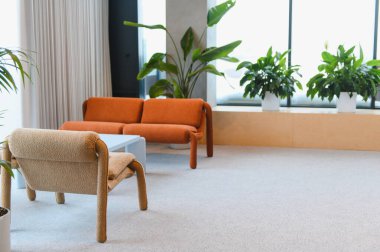 Turuncu bir kanepe, açık bej bir koltuk ve çok sayıda saksı yeşili içeren modern iç mekan, sakin bir atmosfer yaratıyor.