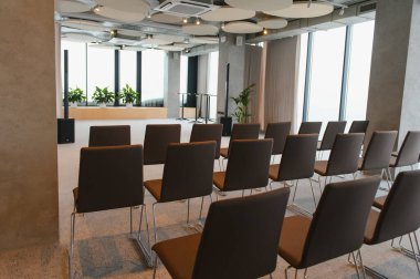 Sahneye bakan sandalyeleri olan, iş toplantıları, seminerler ya da etkinlikler için hazırlanmış modern, boş bir konferans odası. Büyük pencereler doğal ışık sunar