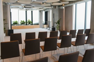 Kürsüye bakan sıra sıra boş sandalyelerle konferans odası, geniş pencereler ve minimalist tasarım