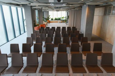 Geniş bir konferans veya seminer odasını dolduran kahverengi sandalyeler, projektör ve geniş pencereler içeriyor