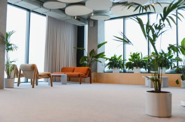 Rahat koltuklar, geniş pencereler ve rahatlatıcı biyofilik atmosfer yaratan sayısız yeşil bitkilerle dolu geniş modern ofis salonu.