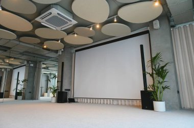 Konferans salonu bir sunum ya da kurumsal etkinlik için hazırlanıyor. Büyük projektör ekran, ses sistemi ve modern tavan tasarımı içerir.