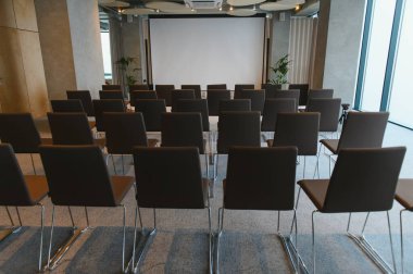 Kurumsal bir etkinlik için kurulmuş çağdaş bir toplantı salonunda boş sandalye dizileri büyük boş bir projektör ekranına bakıyor.