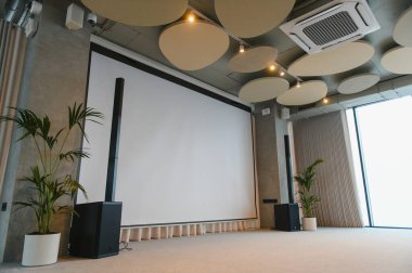 Büyük beyaz ekran ve sütun konuşmacılarının yer aldığı modern sunum salonu, iş toplantıları ve etkinlikler için hazır