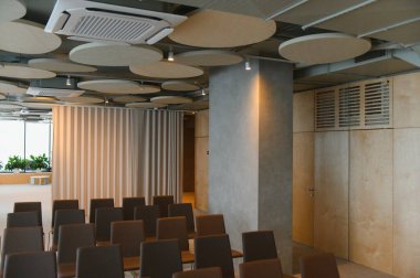 Çağdaş bir oditoryum alanını akustik tavan panelleri ve açık ahşap duvarlarla dolduran kahverengi sandalyeler.