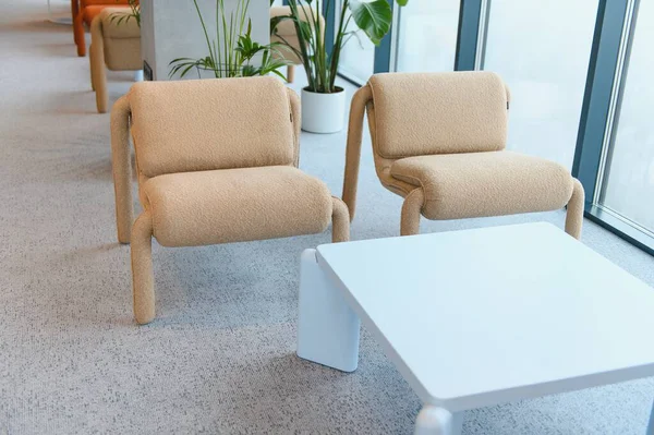 Çağdaş bir ofiste boş bir dinlenme odası, iki şık bej koltuk ve pencere kenarında beyaz bir kahve masası var.