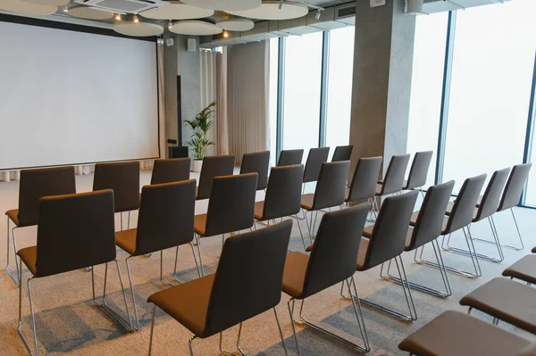 Geniş beyaz projeksiyon ekranı ve parlak pencerelere bakan boş sandalyelerle modern seminer salonu.