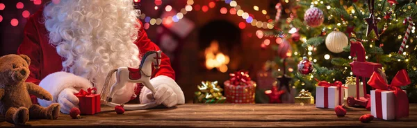 Kerstman zit aan een tafel met geschenken en speelgoed — Stockfoto